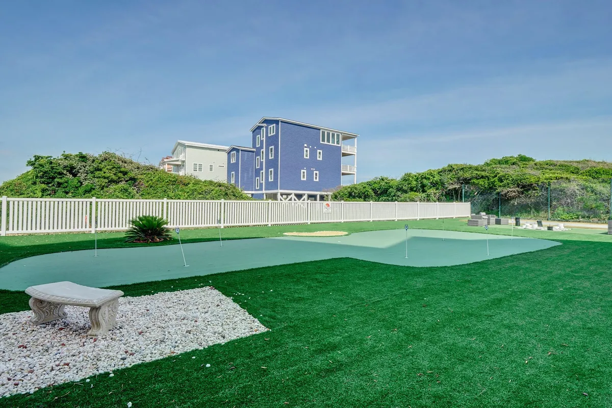 This image displays a lush putting green bordered by green landscaping and a blue house, with a stone bench providing a spot to relax between games. The scene is bright and leisurely, hinting at fun, low-key group activities and peaceful moments outdoors.