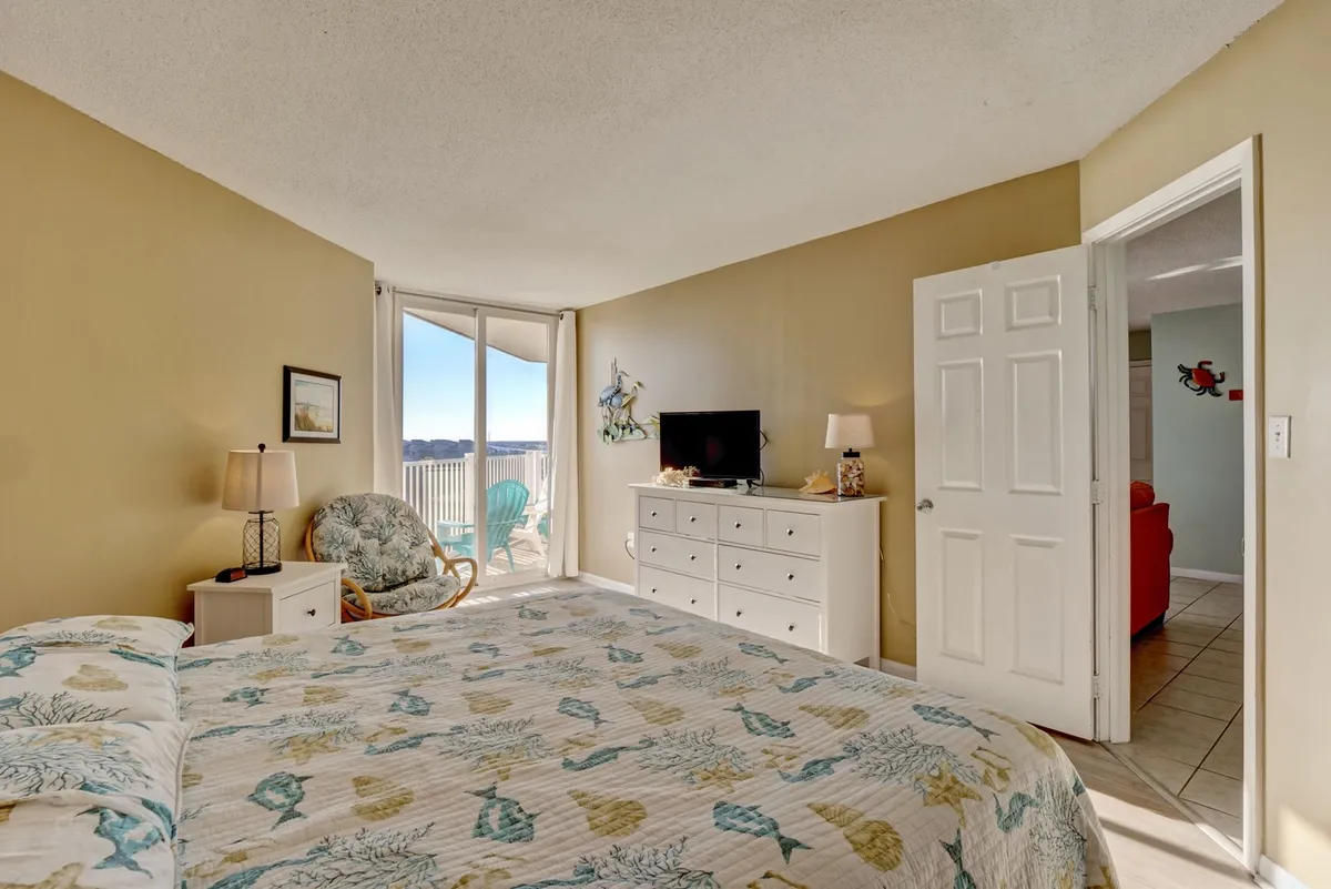 This image showcases a serene bedroom with a bed adorned in a patterned quilt, sliding glass doors that open to a private balcony, a comfortable chair for relaxation, and a dresser with a television. The room is bright, airy, and exudes a tranquil atmosphere, perfect for unwinding after a busy day.