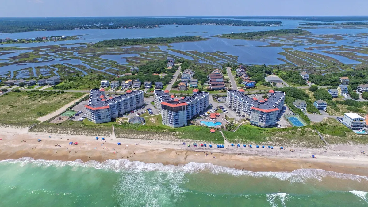 A panoramic exterior view showcasing beachfront properties, a sparkling pool area, colorful umbrellas, marshland, and rolling ocean waves. The lively scene captures the essence of a laid-back seaside resort atmosphere with many opportunities for outdoor exploration and relaxation.