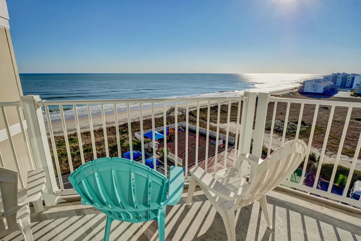 The image captures a sunlit balcony with comfortable chairs overlooking a stunning ocean view. Sunlight glistens on the water, creating a tranquil atmosphere perfect for relaxing, watching dolphins swim by, or stargazing at night. The scene conveys serenity and the quintessential coastal escape.