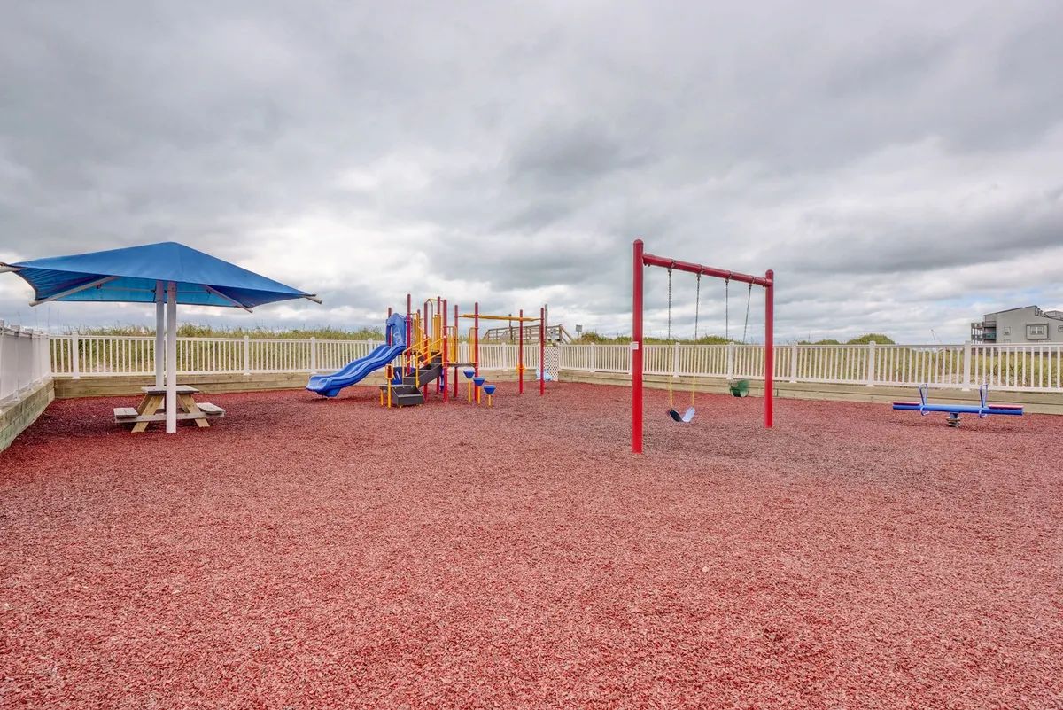 A vibrant outdoor playground with swings, a picnic table, and a sunshade structure sits ready for children’s adventures. Families gather around while kids play energetically in the bright, cheerful environment.