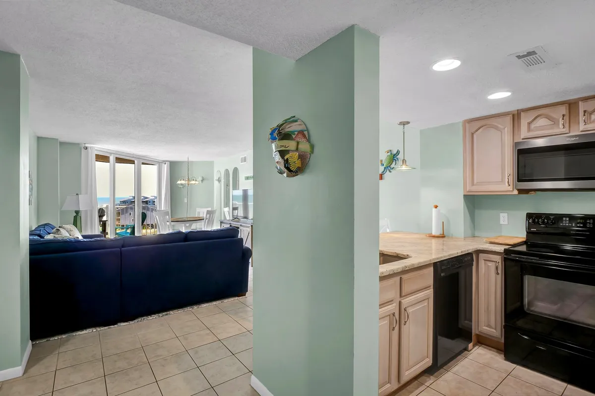This image offers a broad view from the living area into the kitchen, with a blue sofa, dining table, and plenty of natural light. The kitchen is visible in the background, reinforcing the open-concept flow and the kitchen’s integration into the heart of vacation life.