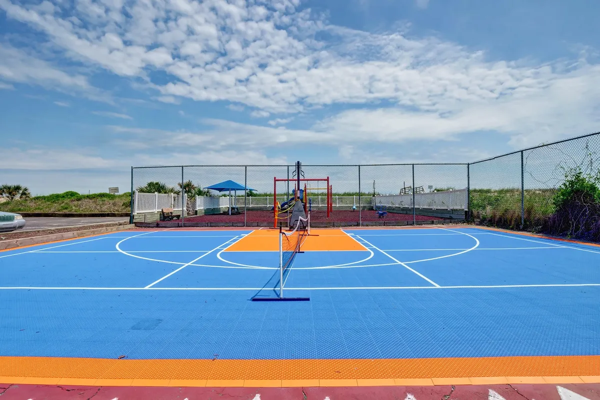 An expansive, multi-use sports court with a basketball hoop, tennis net, and a colorful court surface. In the background, a play area is visible, emphasizing the variety of activities available.