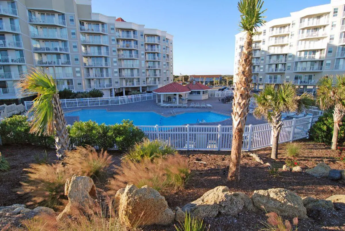 A beautifully landscaped outdoor resort pool surrounded by swaying palm trees and resort buildings. Lounge chairs are set up around the pool for sunbathing, and the area is vibrant and inviting, suggesting a lively social atmosphere.
