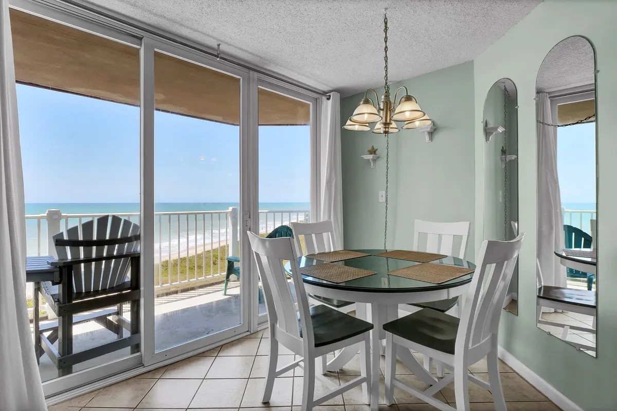 A bright dining area with a table set near large windows that frame a breathtaking view of the Atlantic Ocean. The room is accented by green walls and simple, comfortable furnishings, inviting guests to enjoy meals with the sea as a backdrop.