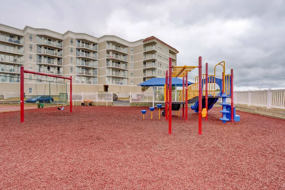 A modern, colorful playground with swings and climbing structures set atop a cheerful red rubber ground cover. Families are gathered nearby, and the building façade creates a safe, welcoming backdrop.