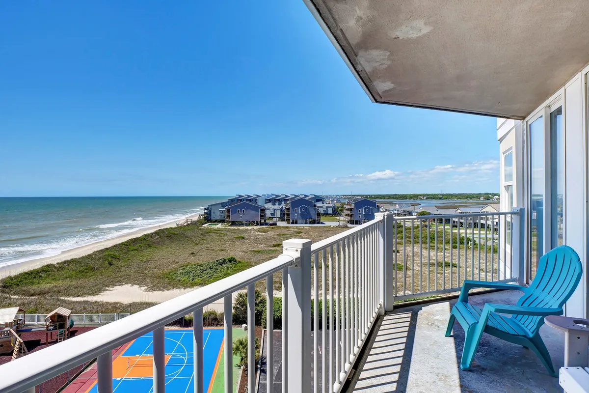 This image showcases an inviting balcony with comfortable seating overlooking a colorful sports court and the vast ocean beyond. The surrounding beach houses, open sky, and visible court surface provide a clear sense of the resort's recreational amenities and beautiful coastal setting. The image captures both opportunities for active play and tranquil moments spent enjoying the view.