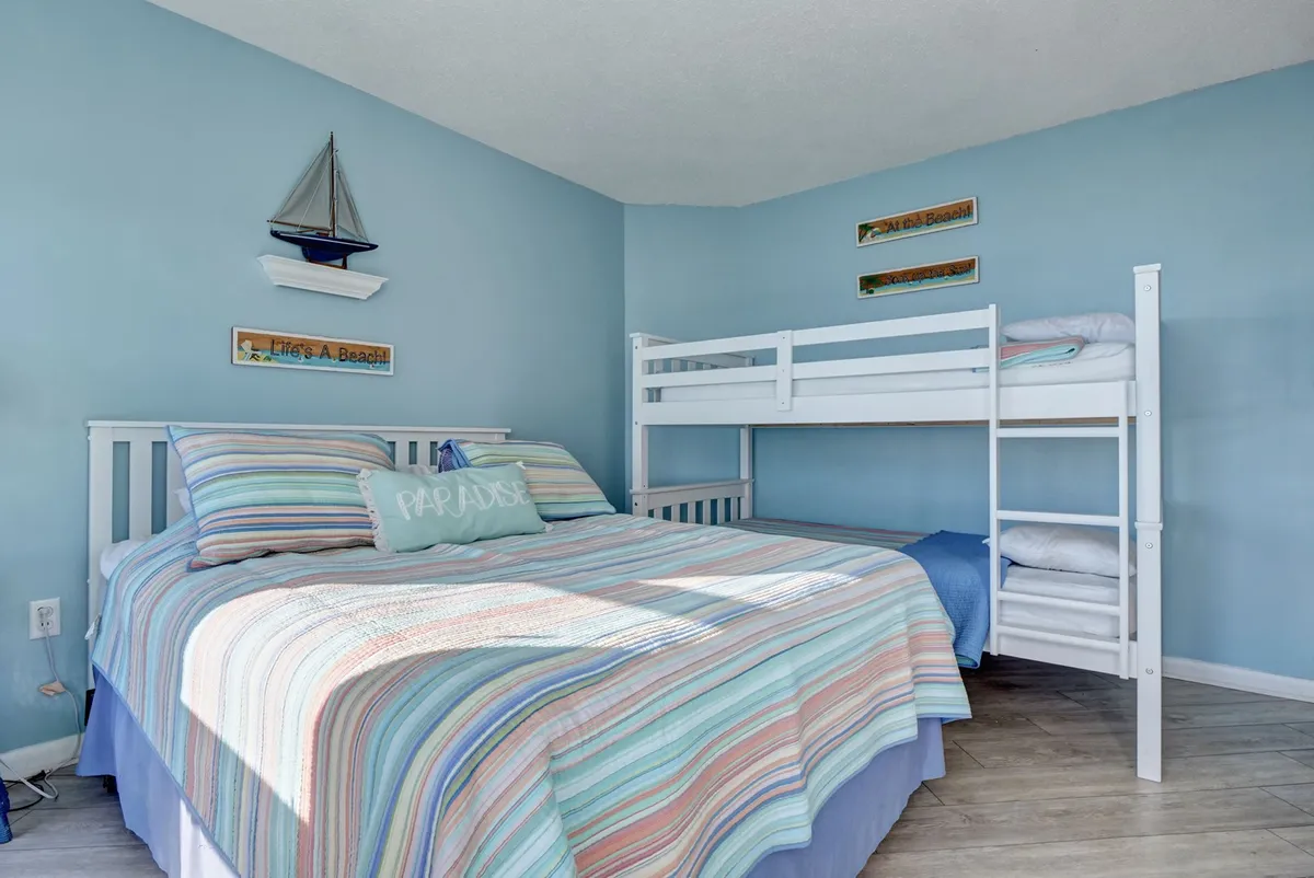 A playful, family-friendly bedroom featuring a queen-size bed and a bunk bed with striped bedding, accented by nautical decor like a decorative sailboat and wall decorations. The room feels inviting and versatile, ideal for families or groups of friends traveling together.