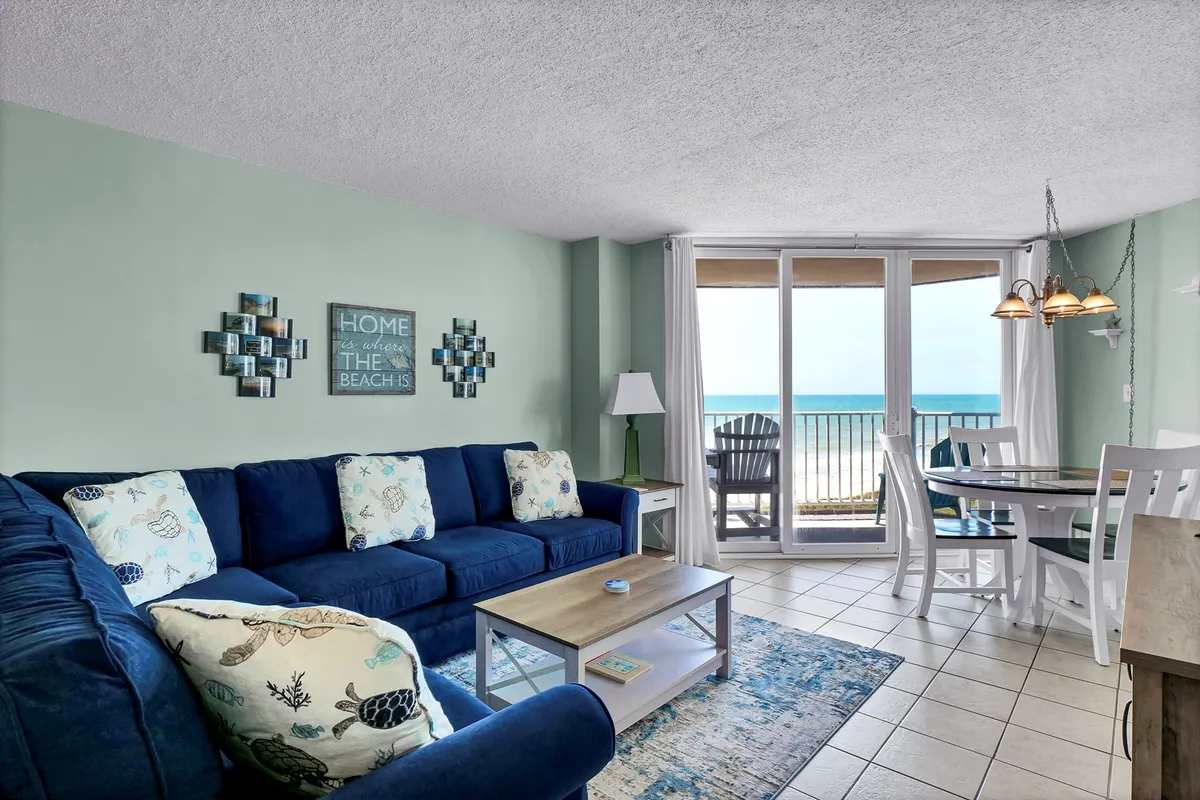 A cozy open-concept living and dining area featuring a blue sofa, dining table, and large glass doors that open to a balcony with sweeping ocean views. Beach-themed wall art adds a coastal touch, and natural light floods the space, highlighting the inviting decor.